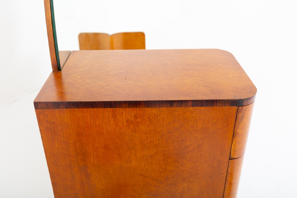 An asymmetric Art Deco dressing table featuring warm birds eye maple veneer, a large circular mirror, a four-drawer stack on the left, and tiered open shelving on the right. View V