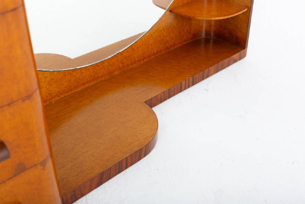 An asymmetric Art Deco dressing table featuring warm birds eye maple veneer, a large circular mirror, a four-drawer stack on the left, and tiered open shelving on the right. View VI