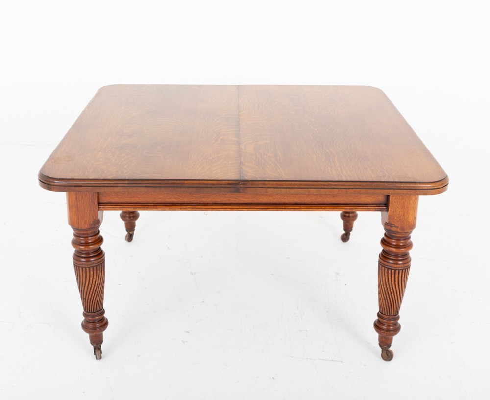 A Victorian blonde oak extending dining table featuring rounded corners, turned spiral-fluted legs, and brass castors against a white background. View II