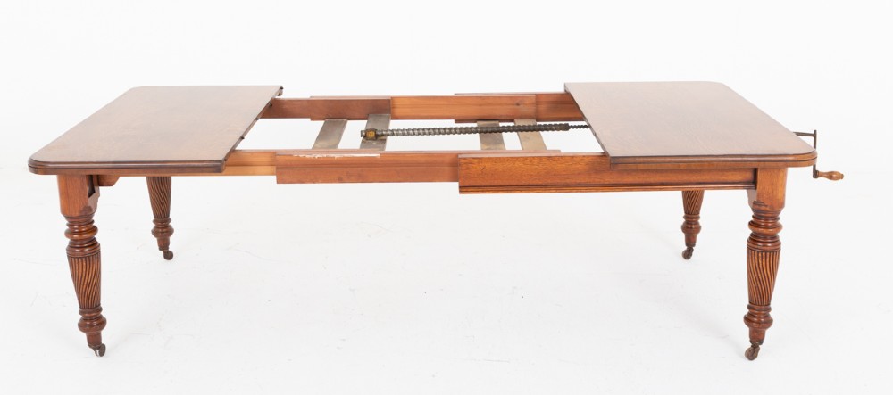 A Victorian blonde oak extending dining table featuring rounded corners, turned spiral-fluted legs, and brass castors against a white background. View V