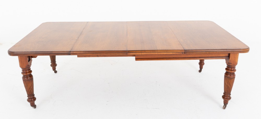 A Victorian blonde oak extending dining table featuring rounded corners, turned spiral-fluted legs, and brass castors against a white background. View VI