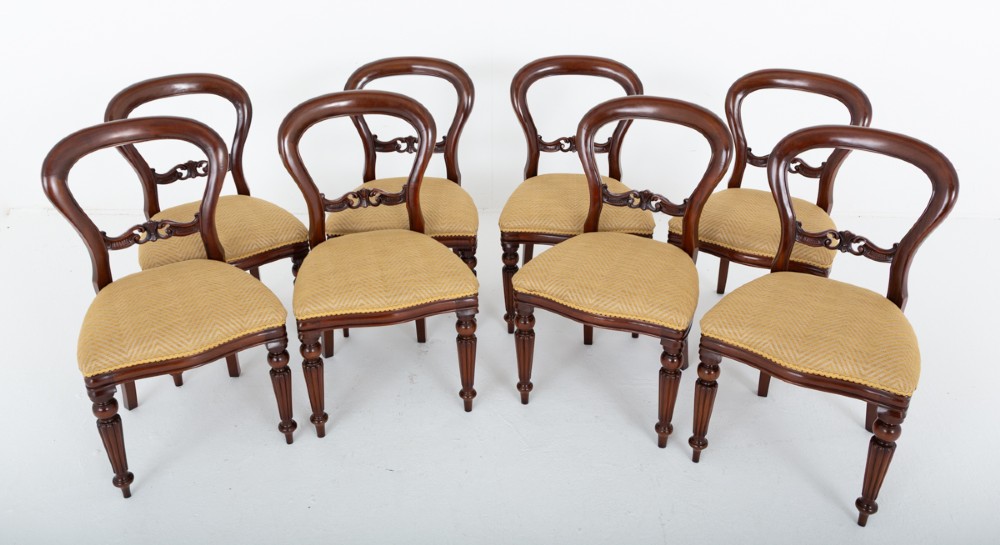 A set of eight Victorian style mahogany dining chairs with rounded balloon backs, carved mid-rails, gold upholstered seats, and fluted front legs.