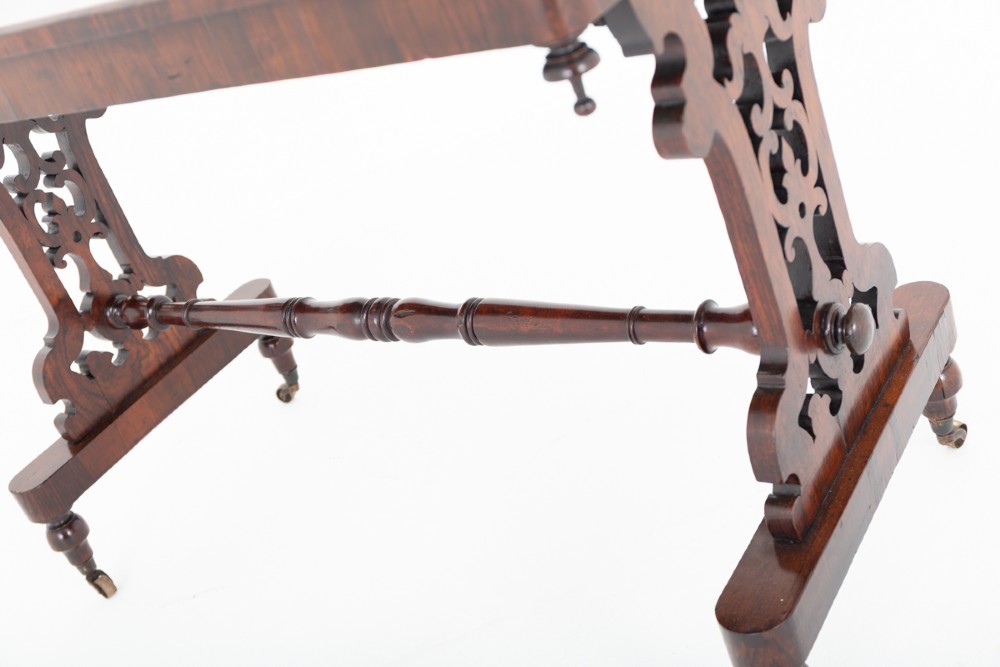A mid-19th century Victorian rosewood stretcher table featuring a rectangular polished top, openwork pierced end supports, a turned central stretcher, and brass castors. View V