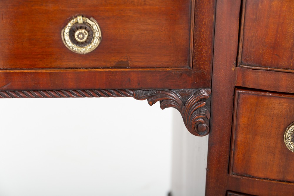 A Georgian Revival mahogany pedestal desk featuring a leather-lined top, curved drawer fronts with brass ring handles, and carved bracket feet. View VI