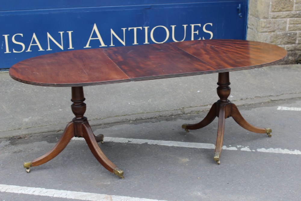 An extendable mahogany twin pedestal dining table featuring rounded ends, reeded edge details, turned central columns, and splayed legs ending in brass castors.