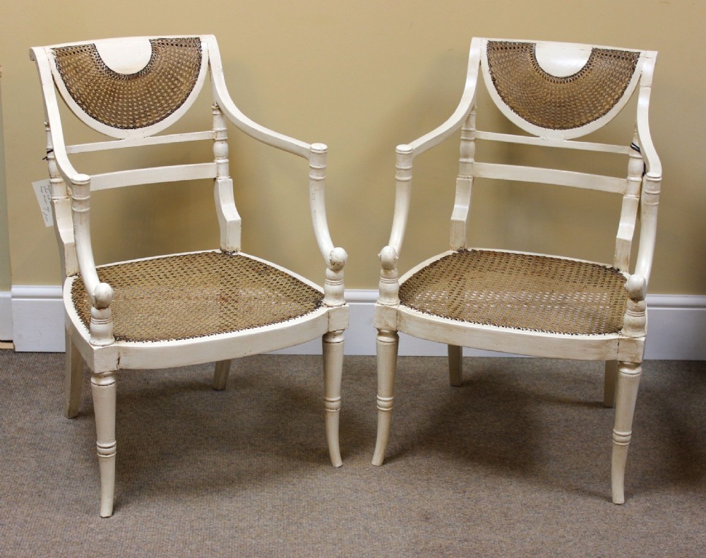 A pair of off-white painted Regency armchairs featuring hand-woven cane seats, crescent caned backrests, and turned front legs.