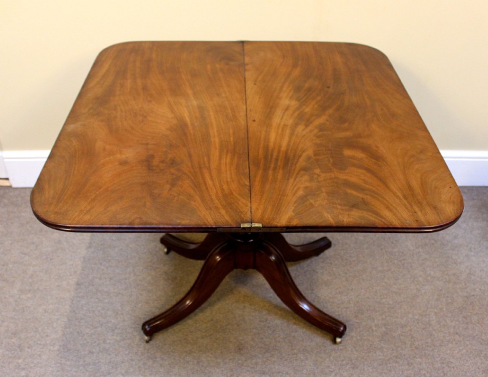 A dark mahogany Regency tea table featuring a rounded fold-over top, a turned central pedestal stem, and four splayed legs with brass castors. View VI