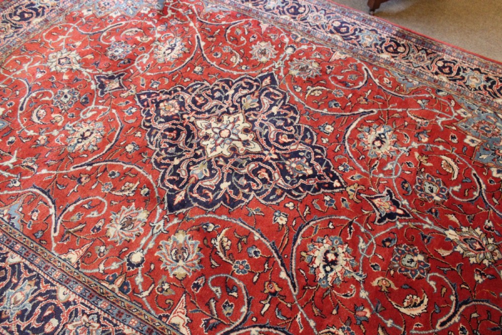 A hand-knotted oriental wool rug with a dark navy central medallion and arabesque scroll motifs set against a red background. View III