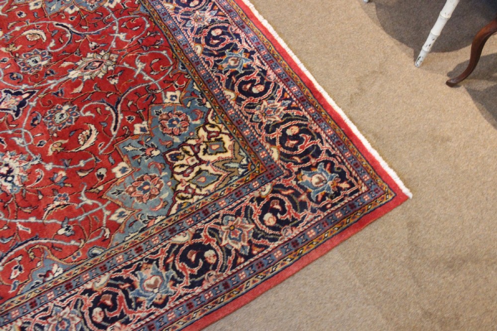 A hand-knotted oriental wool rug with a dark navy central medallion and arabesque scroll motifs set against a red background. View IV