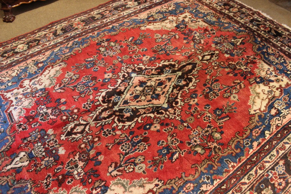 An early 20th-century hand-knotted Persian Sarough rug featuring a red field with a central diamond medallion, floral sprays, and a dark blue border. View VI