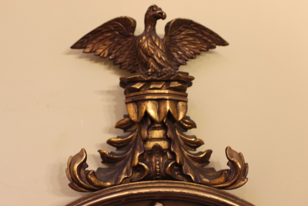 A circular giltwood convex wall mirror topped with a carved eagle cresting and featuring a ball-decorated frame with an acanthus leaf base. View III