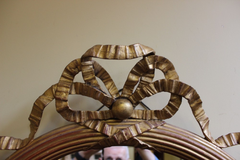 A nineteenth-century oval wall mirror featuring a gilded wood frame carved with a ribbon twist motif and a top bow crest. View III