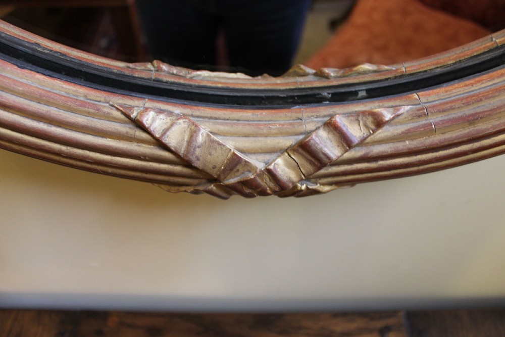 A nineteenth-century oval wall mirror featuring a gilded wood frame carved with a ribbon twist motif and a top bow crest. View IV