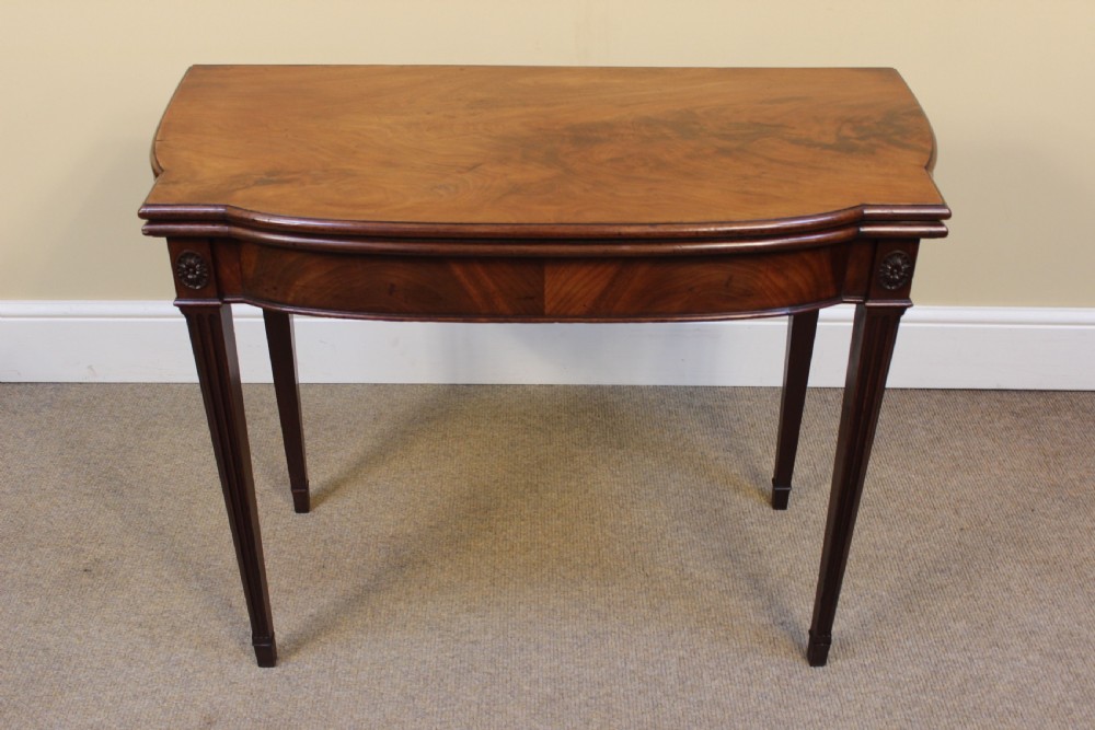 An early 19th-century mahogany fold-over card table featuring a shaped top, flame mahogany veneers, and fluted tapered legs adorned with carved rosettes.