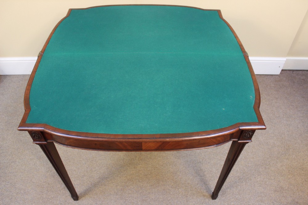 An early 19th-century mahogany fold-over card table featuring a shaped top, flame mahogany veneers, and fluted tapered legs adorned with carved rosettes. View IV
