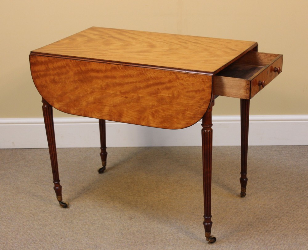 A Regency satinwood Pembroke table with extended drop leaves, a single end drawer, and slender reeded legs resting on brass castors.