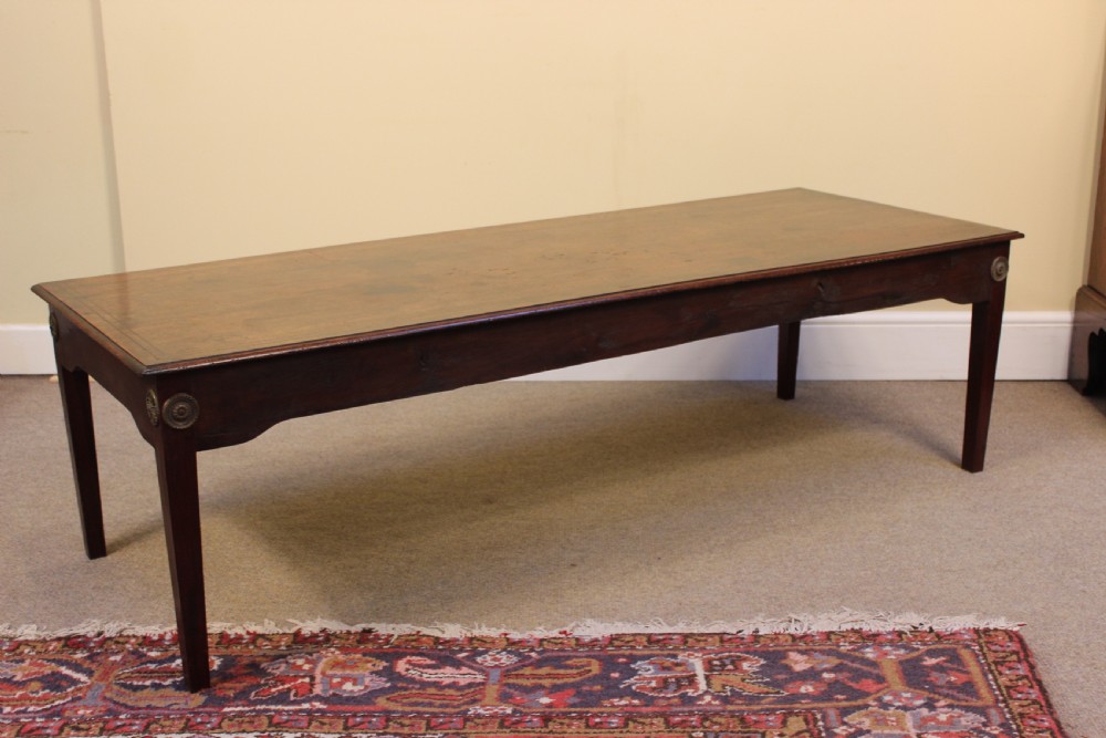 A long rectangular dark mahogany coffee table featuring a polished surface, shaped aprons with circular brass rosette mounts, and four square tapered legs.