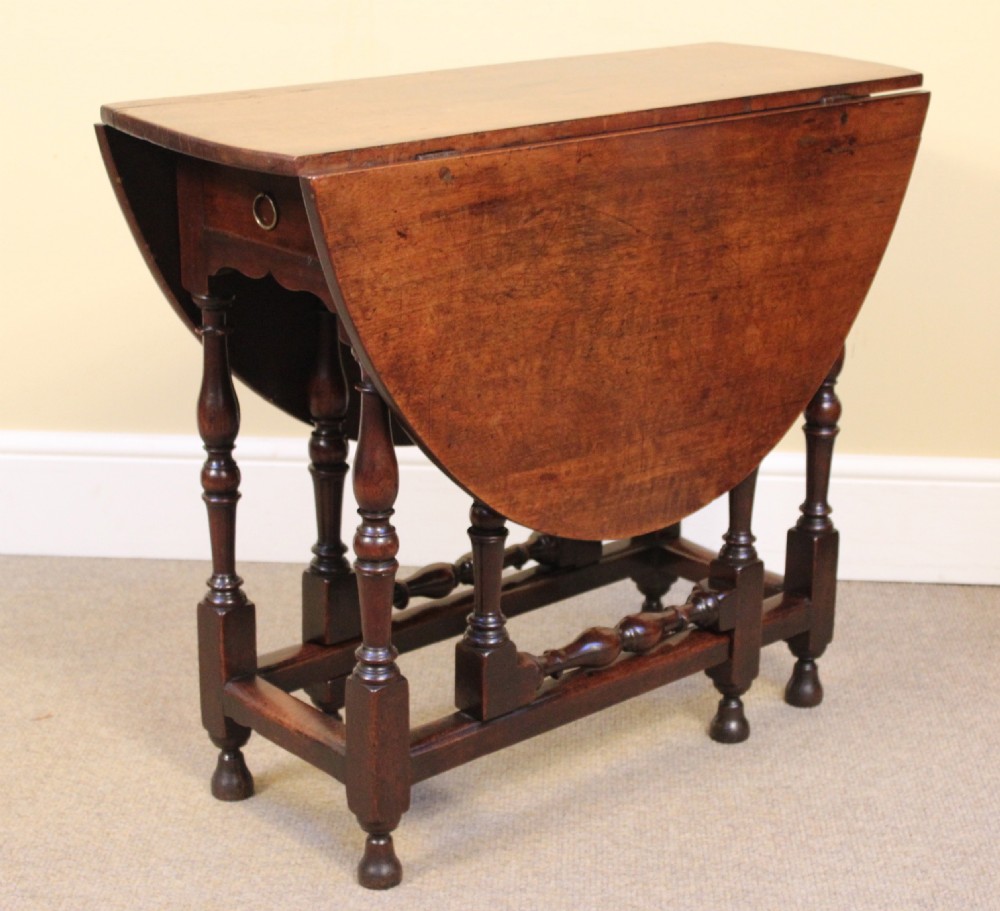 An early eighteenth-century walnut gate leg table featuring baluster-turned legs, a stretcher base, a single drawer, and two drop leaves.