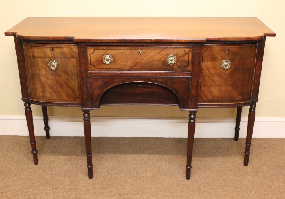 A Regency mahogany breakfront sideboard featuring flame mahogany drawer fronts, brass ring pull handles, a central arched apron, and six turned legs.