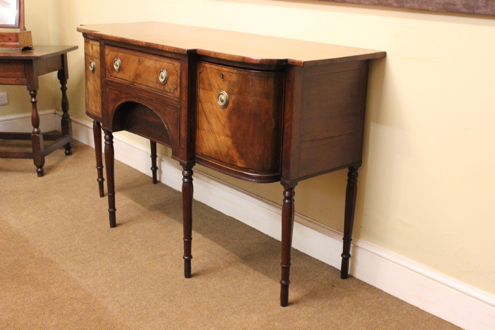 A Regency mahogany breakfront sideboard featuring flame mahogany drawer fronts, brass ring pull handles, a central arched apron, and six turned legs. View II