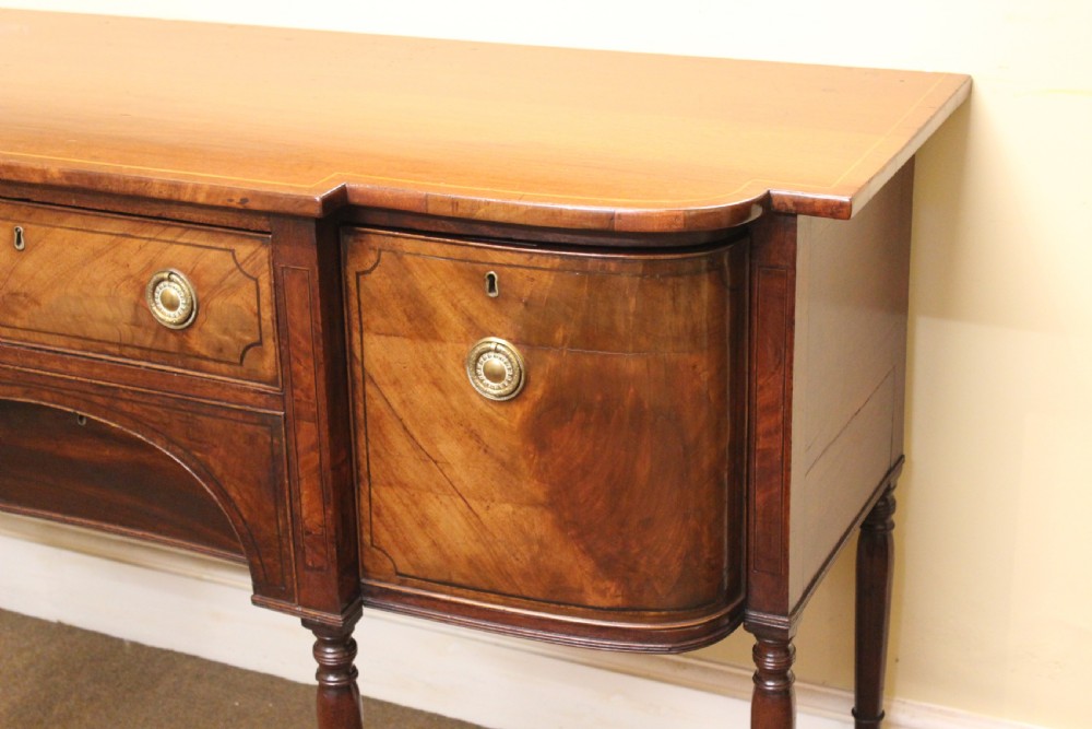 A Regency mahogany breakfront sideboard featuring flame mahogany drawer fronts, brass ring pull handles, a central arched apron, and six turned legs. View V