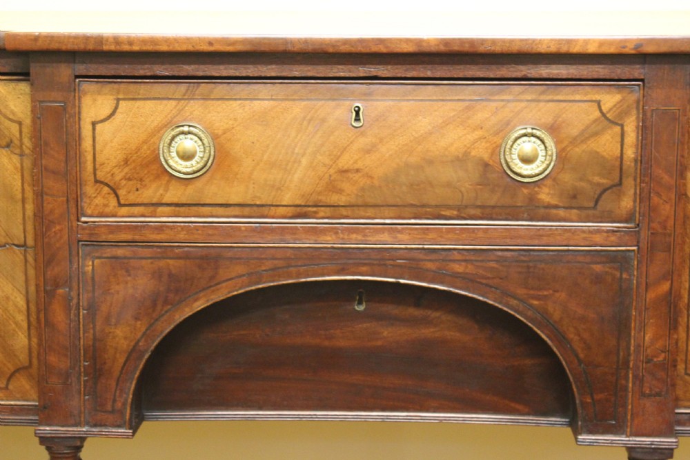 A Regency mahogany breakfront sideboard featuring flame mahogany drawer fronts, brass ring pull handles, a central arched apron, and six turned legs. View VI