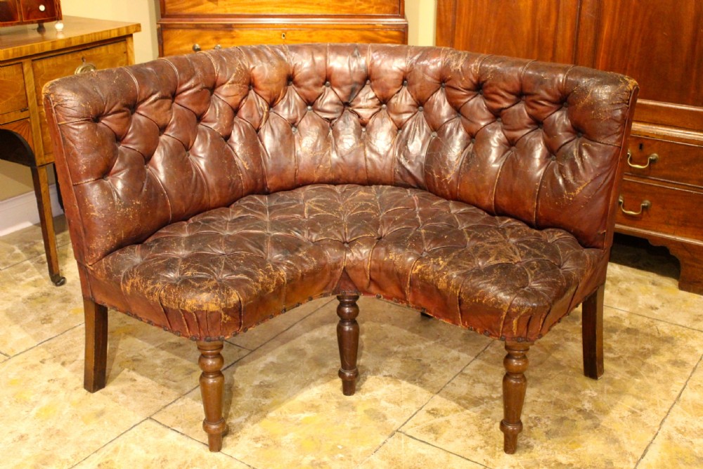 A nineteenth-century curved corner sofa with deep buttoned brown leather upholstery, brass stud details, and turned mahogany legs.