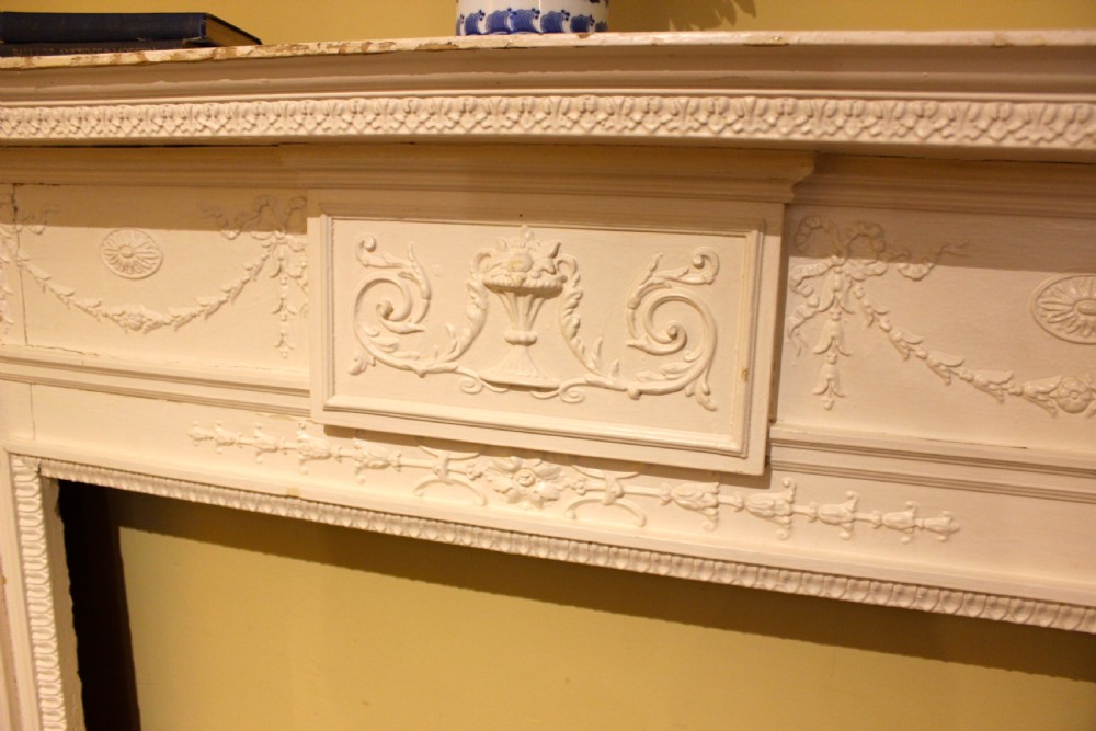 A nineteenth-century white painted pine fire surround featuring carved swag motifs, fluted pilasters, and a central panel decorated with a relief urn. View IV