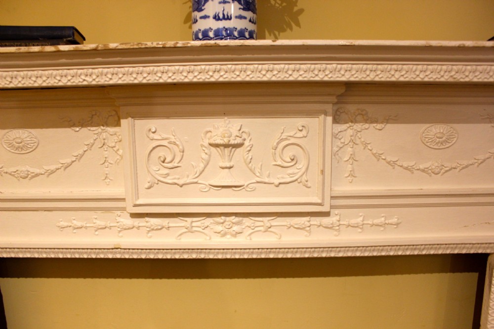 A nineteenth-century white painted pine fire surround featuring carved swag motifs, fluted pilasters, and a central panel decorated with a relief urn. View V