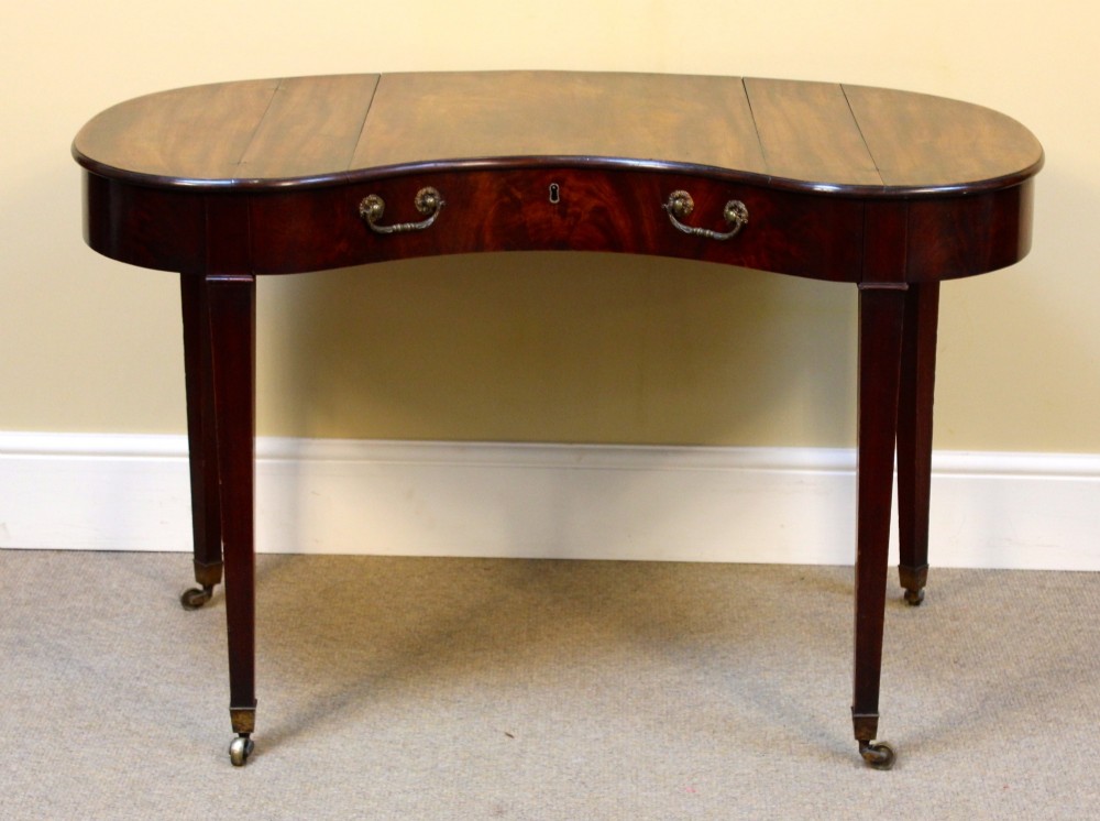 A Georgian mahogany kidney-shaped writing desk featuring tapered legs, brass castors, and a central drawer with brass handles.