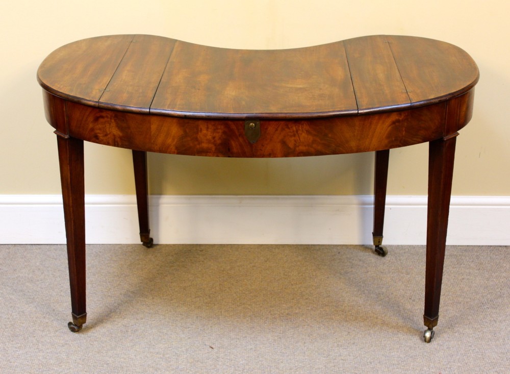 A Georgian mahogany kidney-shaped writing desk featuring tapered legs, brass castors, and a central drawer with brass handles. View V