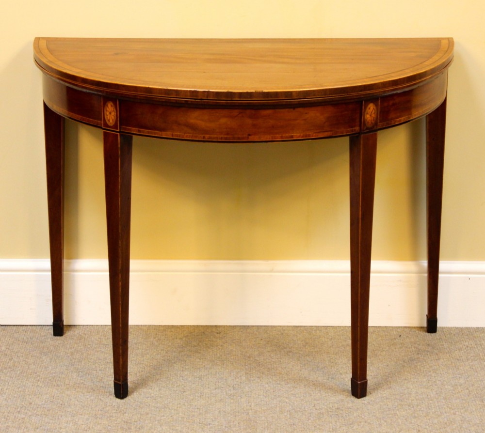 A Regency mahogany demilune card table with a folding top, satinwood stringing, oval patera inlays, and four tapered legs.