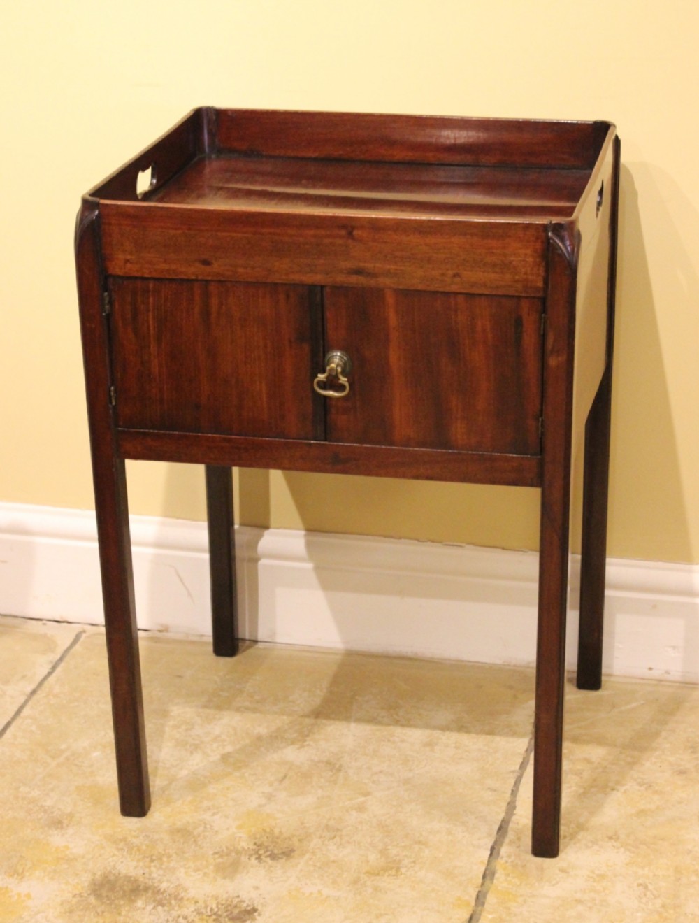 An antique Georgian mahogany bedside table featuring a tray top with cut-out hand-holds, double cupboard doors, a brass pull handle, and straight square legs.
