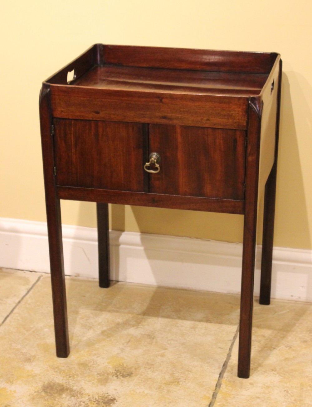 An antique Georgian mahogany bedside table featuring a tray top with cut-out hand-holds, double cupboard doors, a brass pull handle, and straight square legs. View II