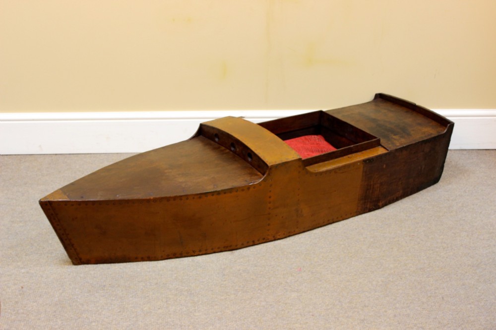 An Edwardian childs model speedboat crafted from brown patinated metal with riveted detailing, dashboard portholes, and a red interior seat.