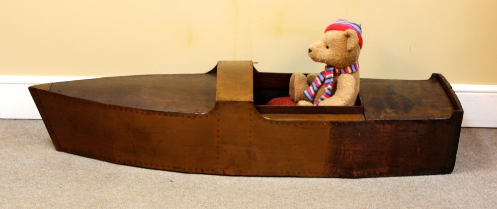 An Edwardian childs model speedboat crafted from brown patinated metal with riveted detailing, dashboard portholes, and a red interior seat. View II