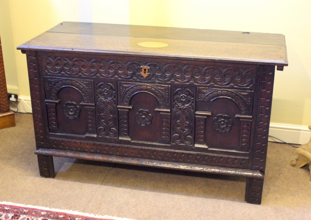 A dark carved oak coffer featuring three arcaded panels with carved floral details and a brass escutcheon.