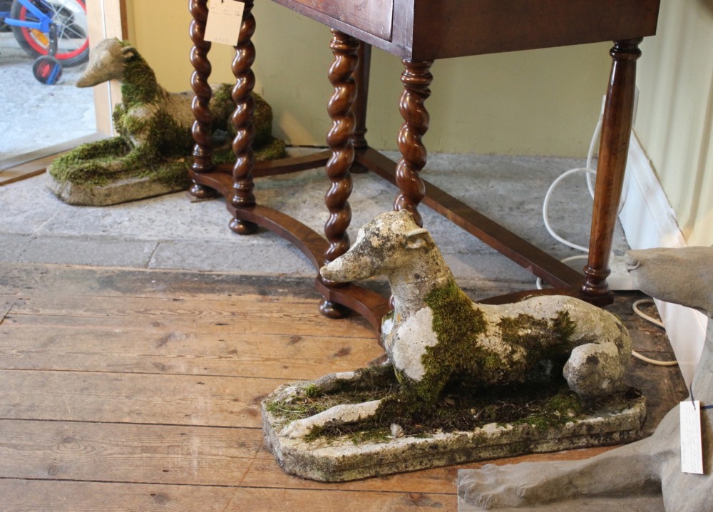 A pair of early 20th-century weathered cast stone statues depicting reclining whippets with natural moss growth on their surface.
