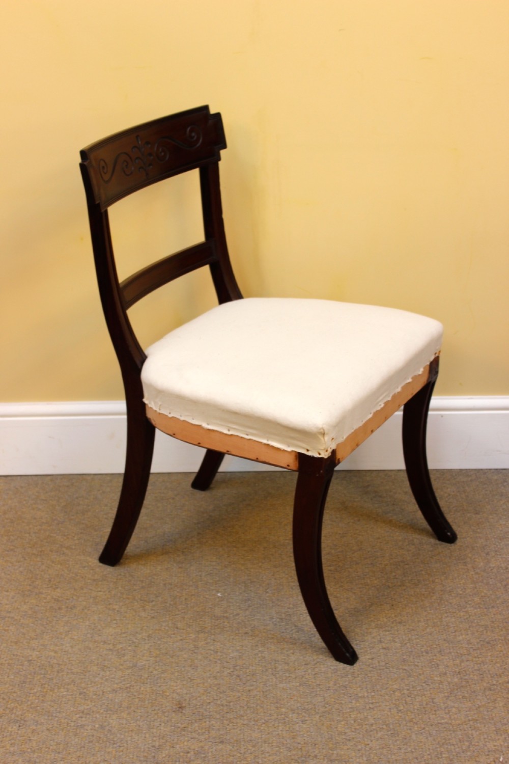 A nineteenth-century mahogany dining chair featuring a carved scroll crest rail, open back, cream upholstered seat, and tapered legs. View VI