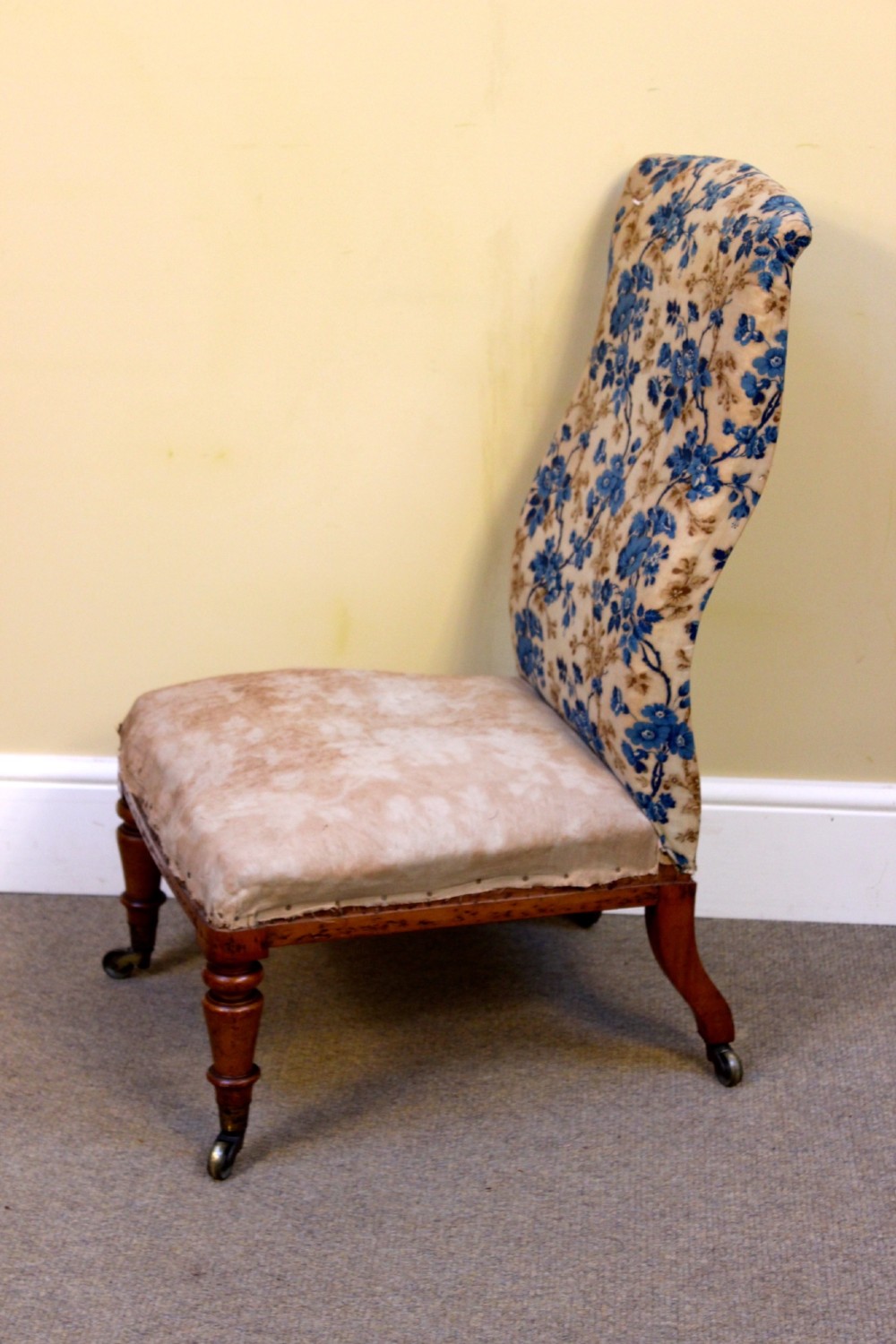 A 19th-century low-seated nursing chair with turned front legs on brass castors, a beige upholstered seat, and a curved backrest covered in blue floral fabric.