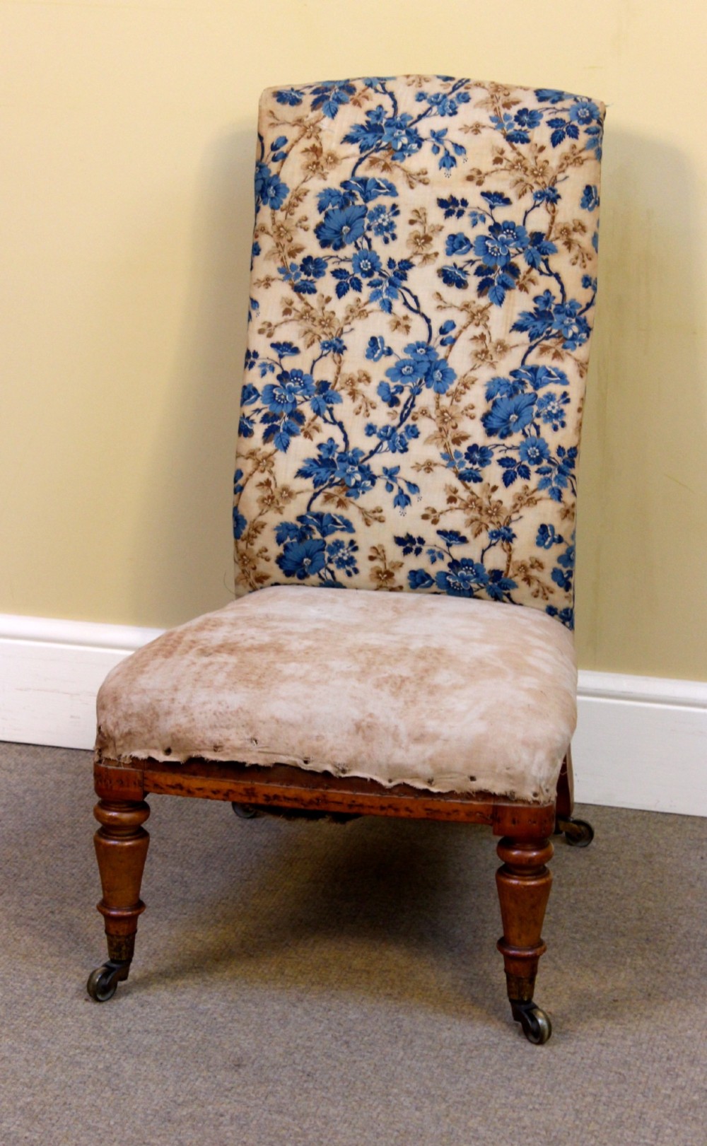 A 19th-century low-seated nursing chair with turned front legs on brass castors, a beige upholstered seat, and a curved backrest covered in blue floral fabric. View II