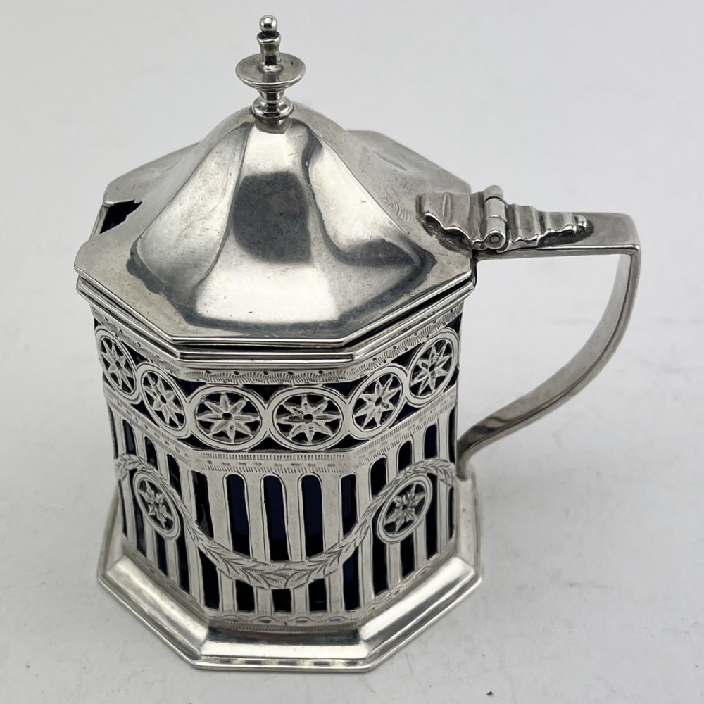 An octagonal Georgian style sterling silver mustard pot featuring pierced fretwork, a hinged lid, and a cobalt blue glass liner.