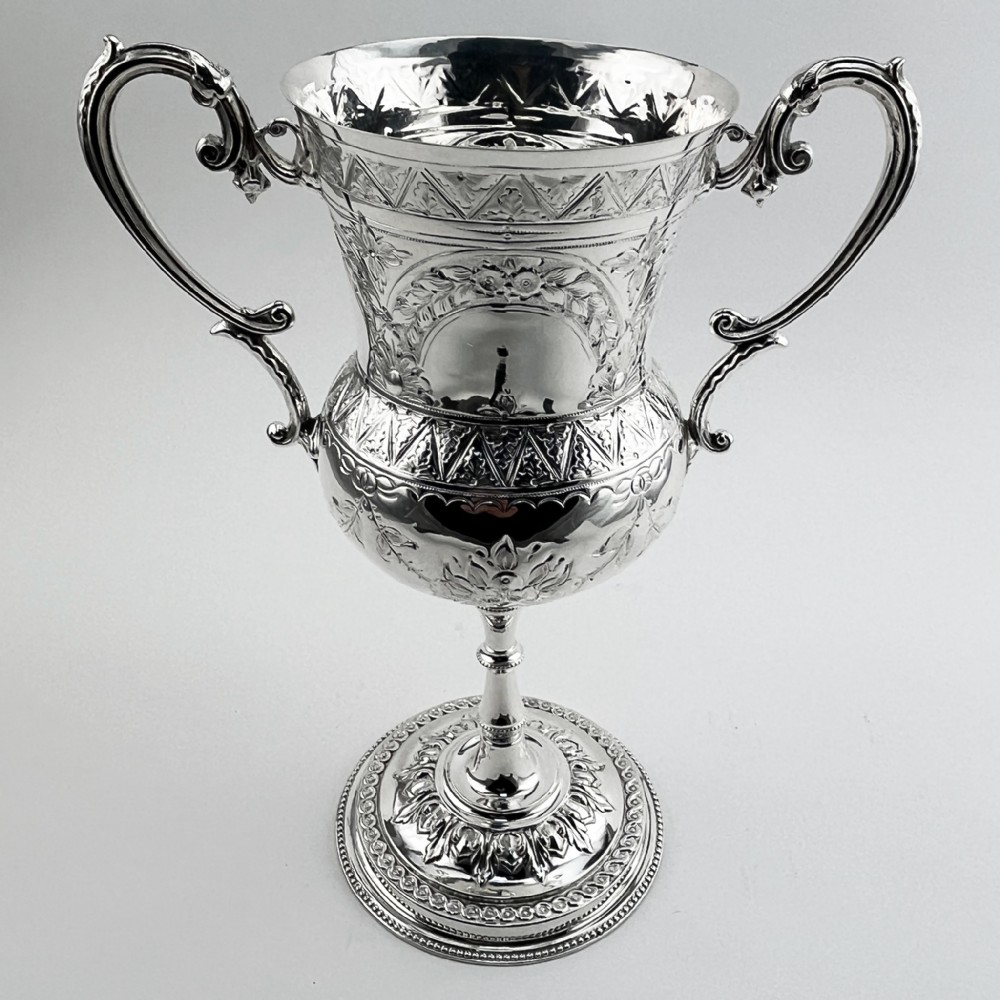 A Victorian silver plated trophy cup featuring ornate scrolled handles, repousse floral engraving, and a circular decorated pedestal foot.