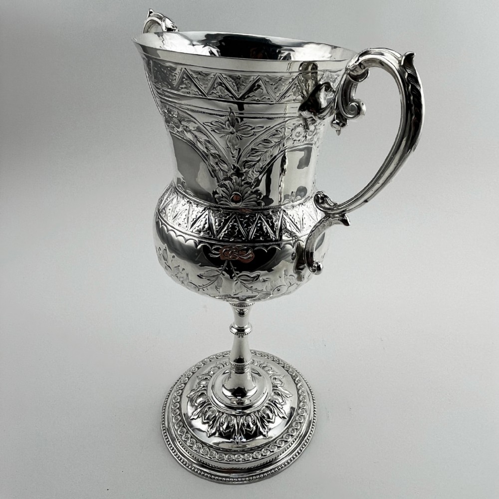 A Victorian silver plated trophy cup featuring ornate scrolled handles, repousse floral engraving, and a circular decorated pedestal foot. View II