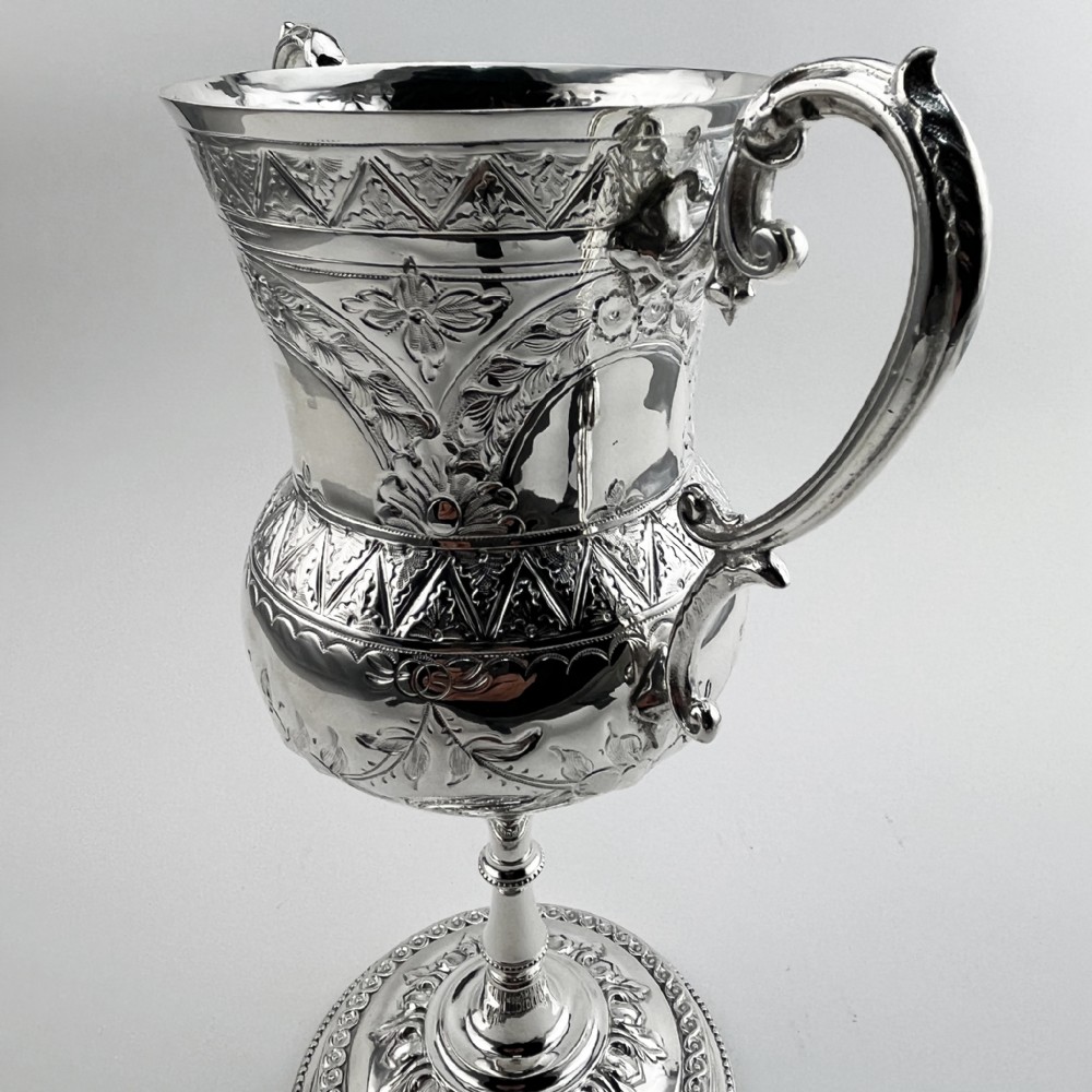 A Victorian silver plated trophy cup featuring ornate scrolled handles, repousse floral engraving, and a circular decorated pedestal foot. View III