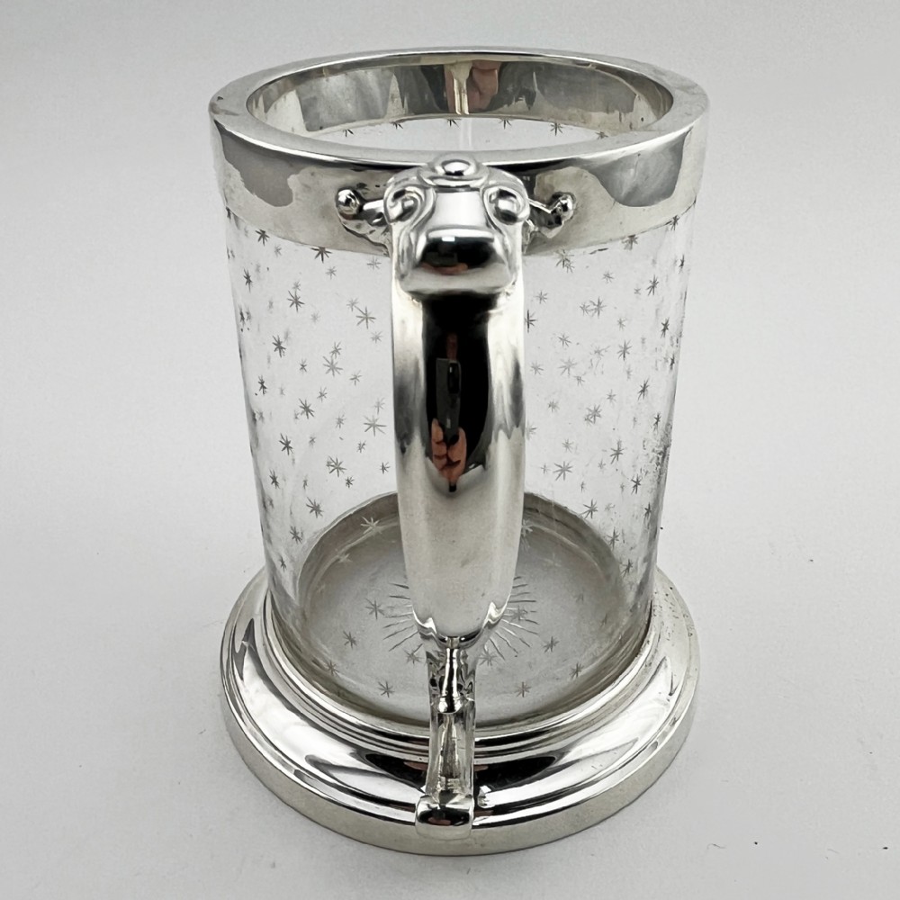 A Victorian clear glass mug featuring etched star motifs, a silver-plated rim, a stepped silver base, and a decorative scrolling handle. View V