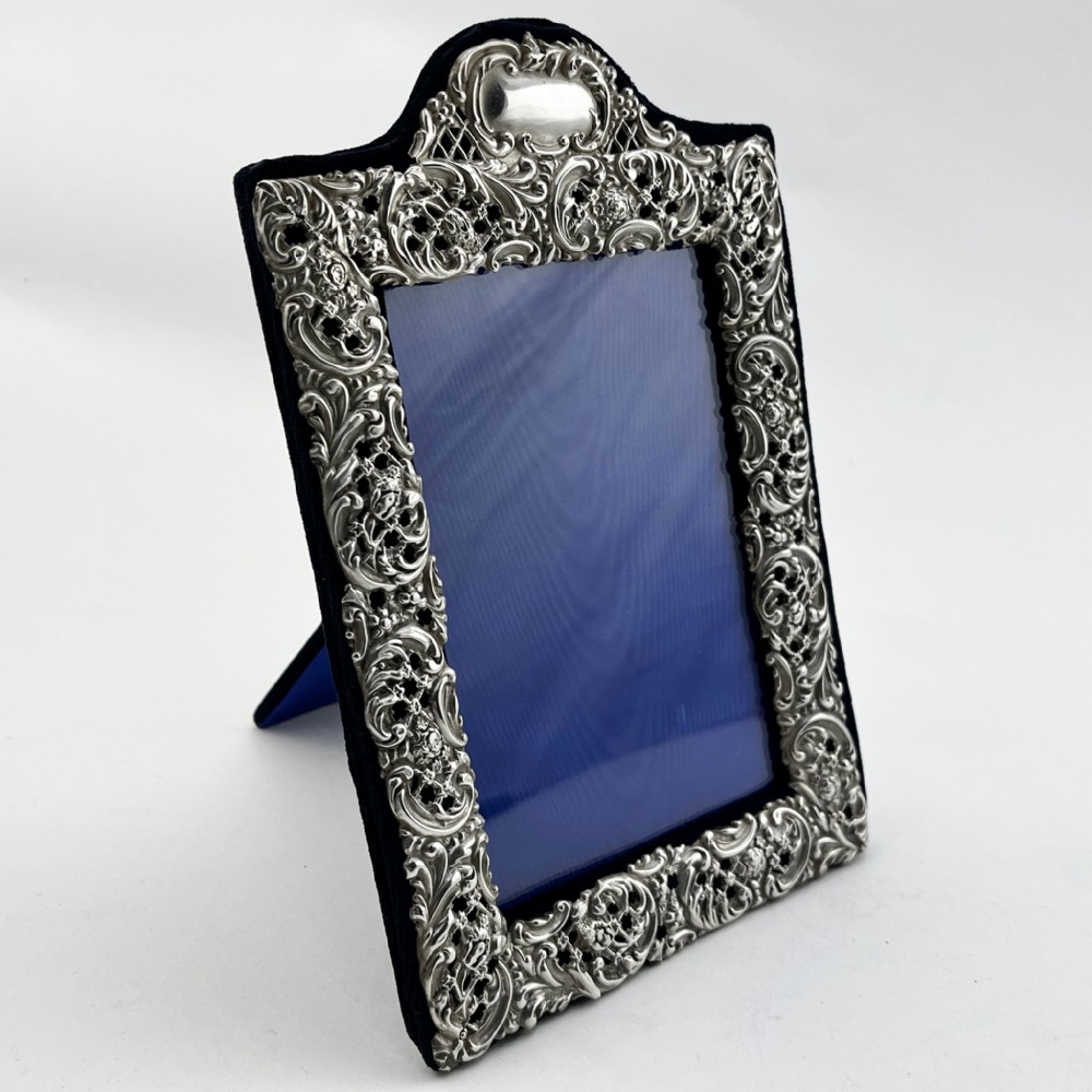 A rectangular sterling silver photo frame featuring intricate floral and scroll repousse metalwork mounted on a dark blue velvet easel backing.