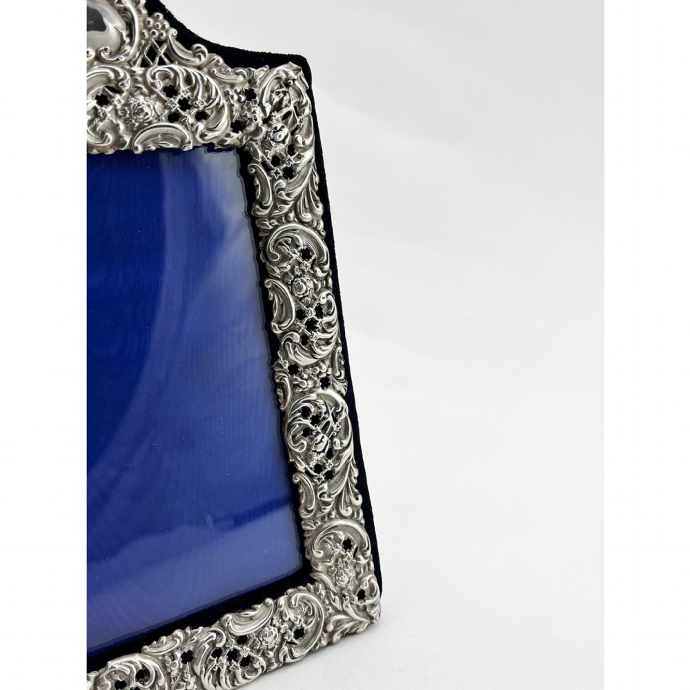 A rectangular sterling silver photo frame featuring intricate floral and scroll repousse metalwork mounted on a dark blue velvet easel backing. View II