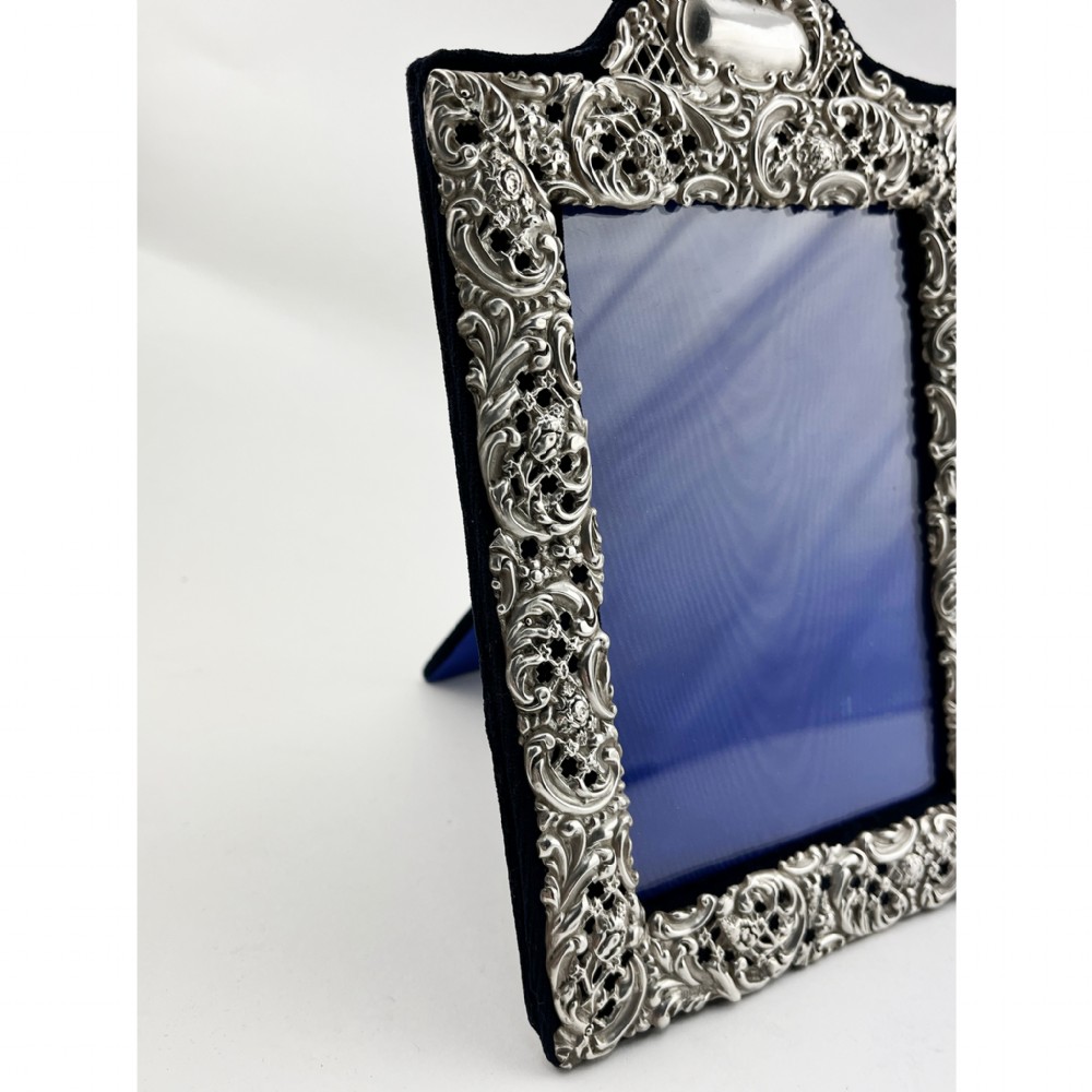 A rectangular sterling silver photo frame featuring intricate floral and scroll repousse metalwork mounted on a dark blue velvet easel backing. View III