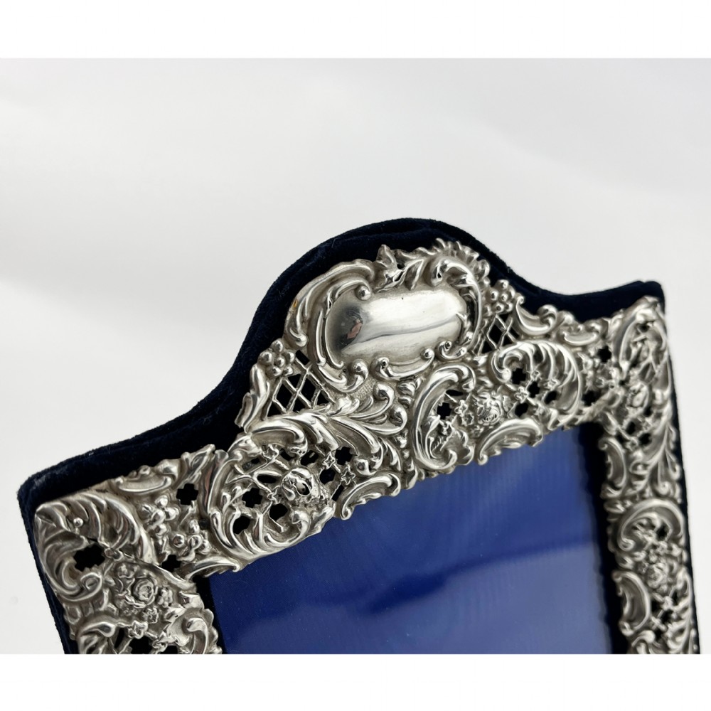 A rectangular sterling silver photo frame featuring intricate floral and scroll repousse metalwork mounted on a dark blue velvet easel backing. View IV